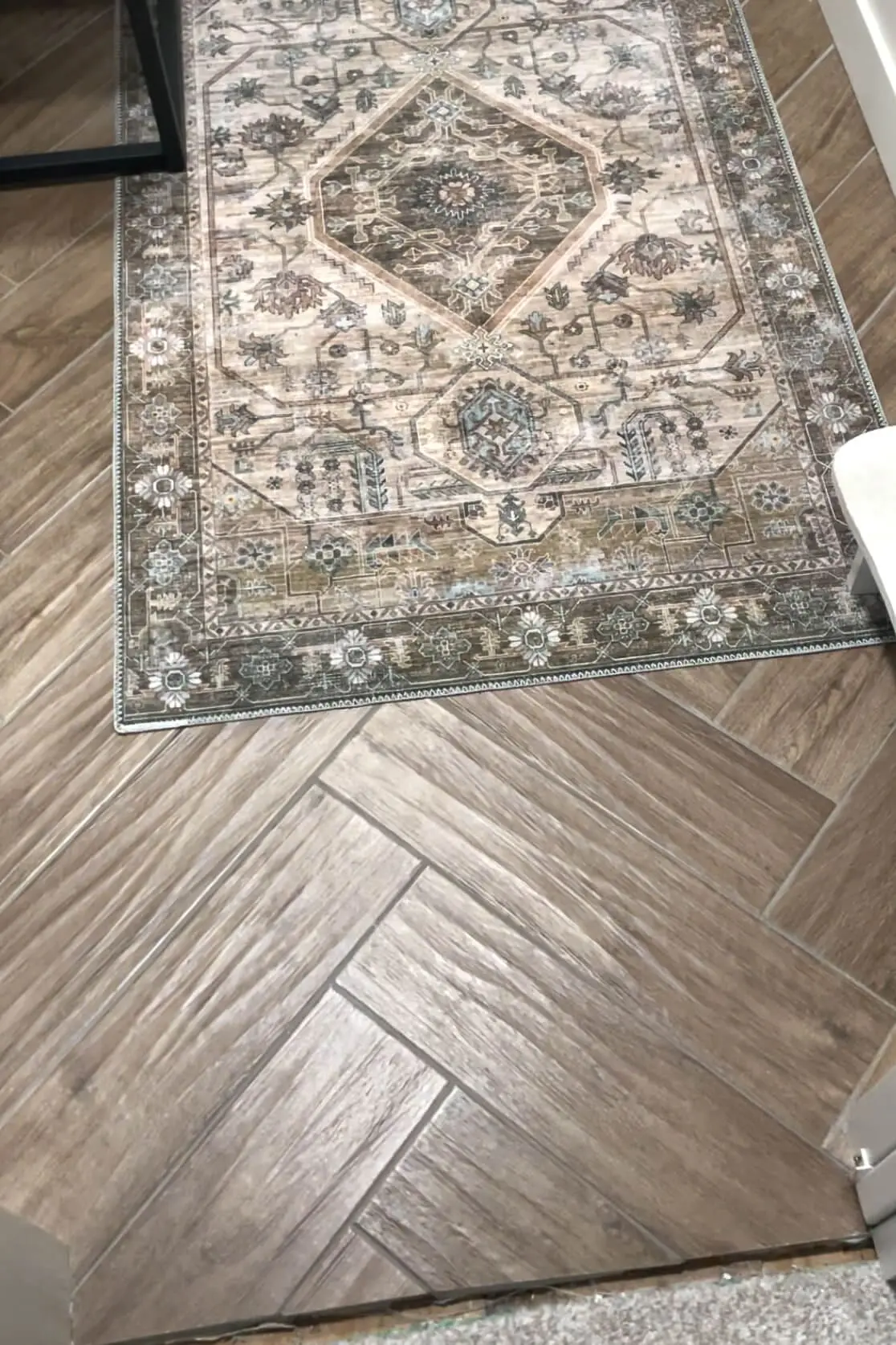 A detailed view of herringbone-patterned wood-look tile flooring, heated by a DIY radiant floor heating system, paired with a vintage-inspired rug for a cozy bathroom upgrade.